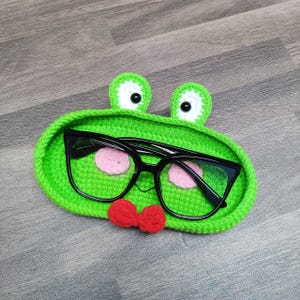 May include: Green crocheted frog-shaped glasses holder with black framed glasses inside and a red bow tie.