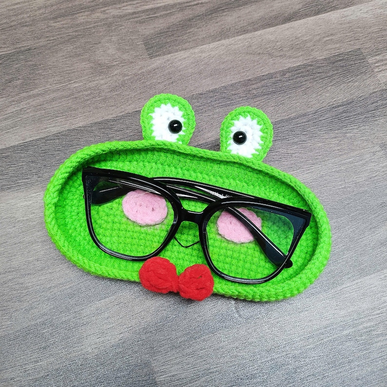 May include: Green crocheted frog-shaped glasses holder with black framed glasses inside and a red bow tie.