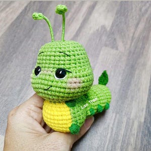 Può includere: Un giocattolo a forma di bruco all'uncinetto fatto a mano, verde e giallo, con grandi occhi neri e un viso sorridente. Il bruco ha antenne verdi e una piccola coda verde. Il giocattolo è tenuto in una mano.