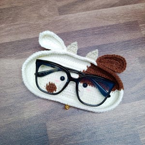 May include: A crocheted cow-shaped glasses holder with brown and white spots. The holder has a gold bell charm and is holding a pair of black-framed glasses.