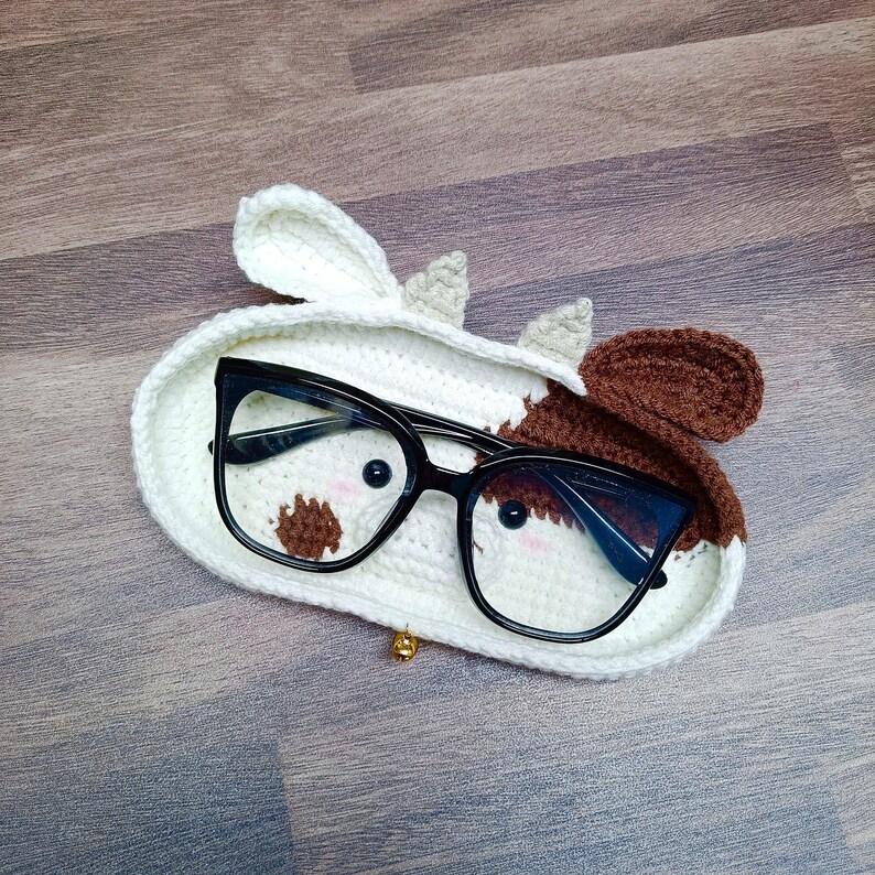 May include: A crocheted cow-shaped glasses holder with brown and white spots. The holder has a gold bell charm and is holding a pair of black-framed glasses.