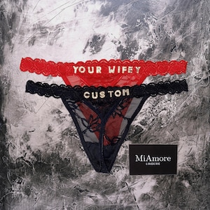 Peut inclure: Deux sous-vêtements de style string. L'un est rouge avec le texte "YOUR WIFEY" en lettres dorées. L'autre est noir avec le texte "CUSTOM" en lettres dorées. Les deux ont une bordure en dentelle. Le logo MiAmore Lingerie est en bas à droite.