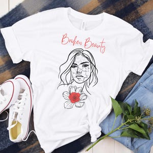 Può includere: Maglietta bianca con un disegno a linea nera di una donna con un fiore rosso sul petto. Il testo "Broken Beauty" è scritto in corsivo rosso sopra la testa della donna.