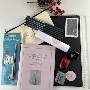 Op de afbeelding: Een assortiment kunstbenodigdheden, waaronder potloden, een kalligrafieset, een schetsboek en een liniaal. Er is ook een boek met de titel "Learn How to Draw in Six Weeks". De items zijn gerangschikt met decoratieve dennenappels en rode bessen.