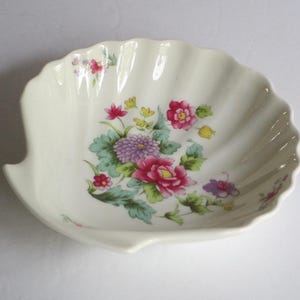 May include: A cream-colored ceramic dish shaped like a seashell with a scalloped edge. The interior features a colorful floral pattern with pink, purple, and yellow flowers surrounded by green leaves, painted in the center and along the rim.