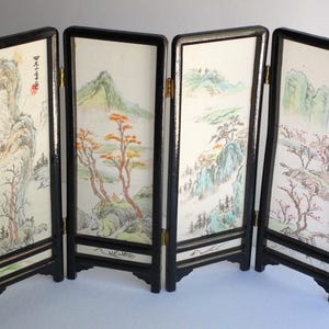 May include: A four-panel folding screen with a black lacquered frame. Each panel features a light-colored insert with a delicate landscape painting in muted green, brown, and red tones. The first panel includes red calligraphy and a red seal mark.