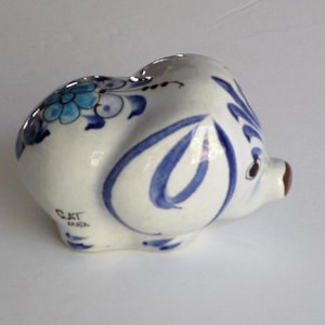 May include: A white ceramic piggy bank featuring hand-painted blue floral patterns and swirling designs. The pig has a small brown snout, dark eyes, and the text "CAT MADA" printed in black on its side. It is a rounded, glossy figurine.