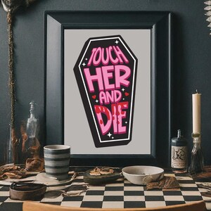 May include: A framed print featuring a coffin shape with the words "TOUCH HER AND DIE" in pink and white lettering. The artwork has a black border and is set against a gray background. The print is displayed on a checkered surface.