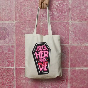 May include: A cream-colored tote bag with long handles. The bag features a black coffin-shaped graphic with the words "TOUCH HER AND DIE" in pink and white lettering. The bag is held up by a hand.