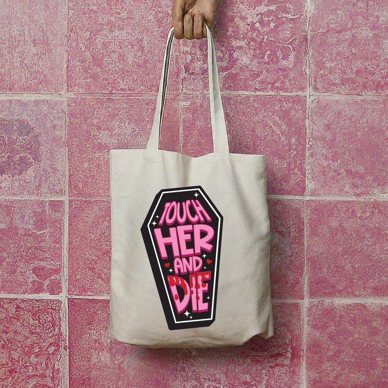 May include: A cream-colored tote bag with long handles. The bag features a black coffin-shaped graphic with the words "TOUCH HER AND DIE" in pink and white lettering. The bag is held up by a hand.