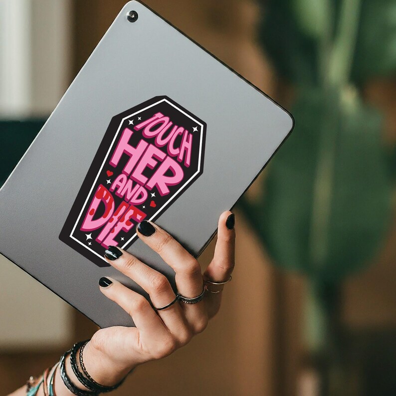 May include: A coffin-shaped sticker with the text "Touch Her and Die" in pink and white lettering. The sticker is on a gray tablet. The image is of a hand holding the tablet.