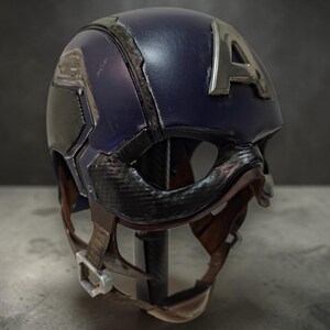 May include: A navy blue Captain America helmet with a silver "A" emblem. The helmet has a black visor and chin strap with a brown leather-like texture. The helmet is displayed on a dark background.