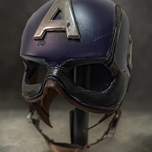 K&ouml;nnte beinhalten: Blauer Captain America Helm mit silbernem "A" auf der Vorderseite. Der Helm hat ein schwarzes Visier und einen Kinnriemen. Eine Nachbildung des Helms der Comicfigur.