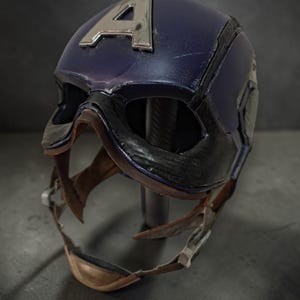 K&ouml;nnte beinhalten: Blau-schwarzer Captain America Helm mit silbernem "A" Emblem. Der Helm hat ein abgenutztes Finish mit sichtbaren Gebrauchsspuren. Der Kinnriemen ist hellbraun und der Helm ist auf einem Metallst&auml;nder montiert.