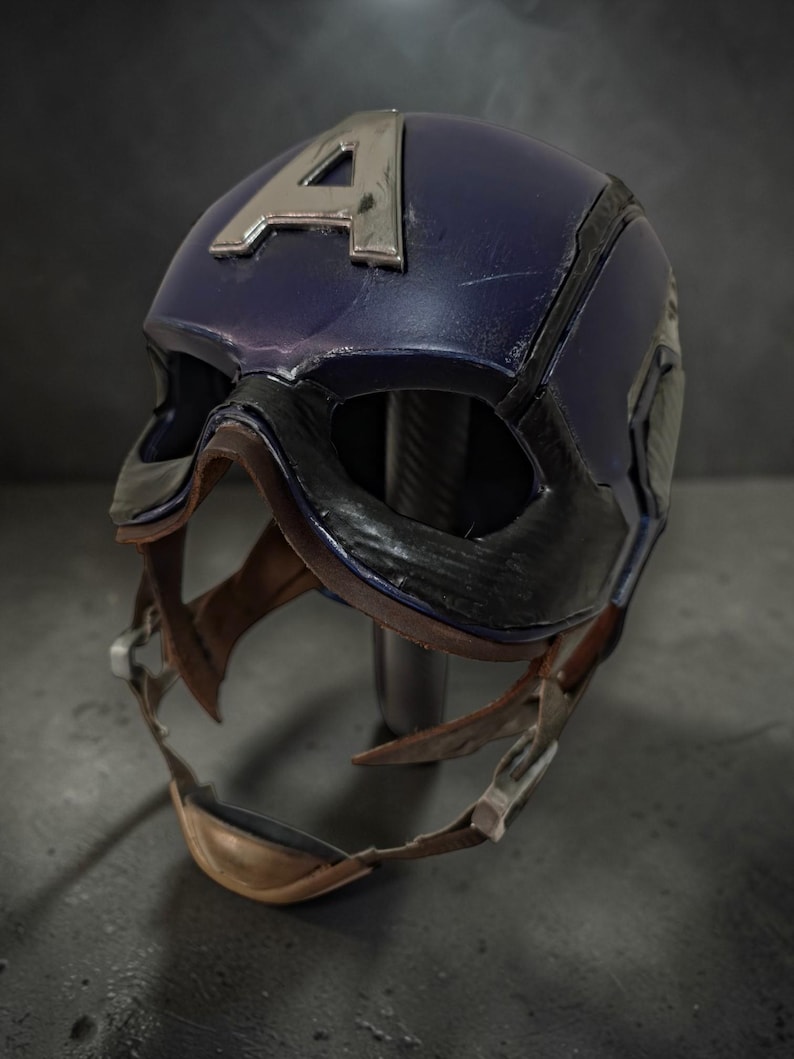 K&ouml;nnte beinhalten: Blau-schwarzer Captain America Helm mit silbernem "A" Emblem. Der Helm hat ein abgenutztes Finish mit sichtbaren Gebrauchsspuren. Der Kinnriemen ist hellbraun und der Helm ist auf einem Metallst&auml;nder montiert.