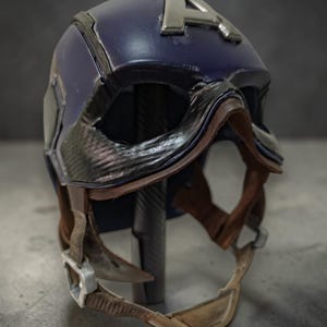 K&ouml;nnte beinhalten: Ein dunkelblauer Helm mit silbernem "A"-Emblem. Der Helm hat ein lederartiges Visier und einen Kinnriemen mit silberner Schnalle. Das Design erinnert an einen Superheldenhelm.
