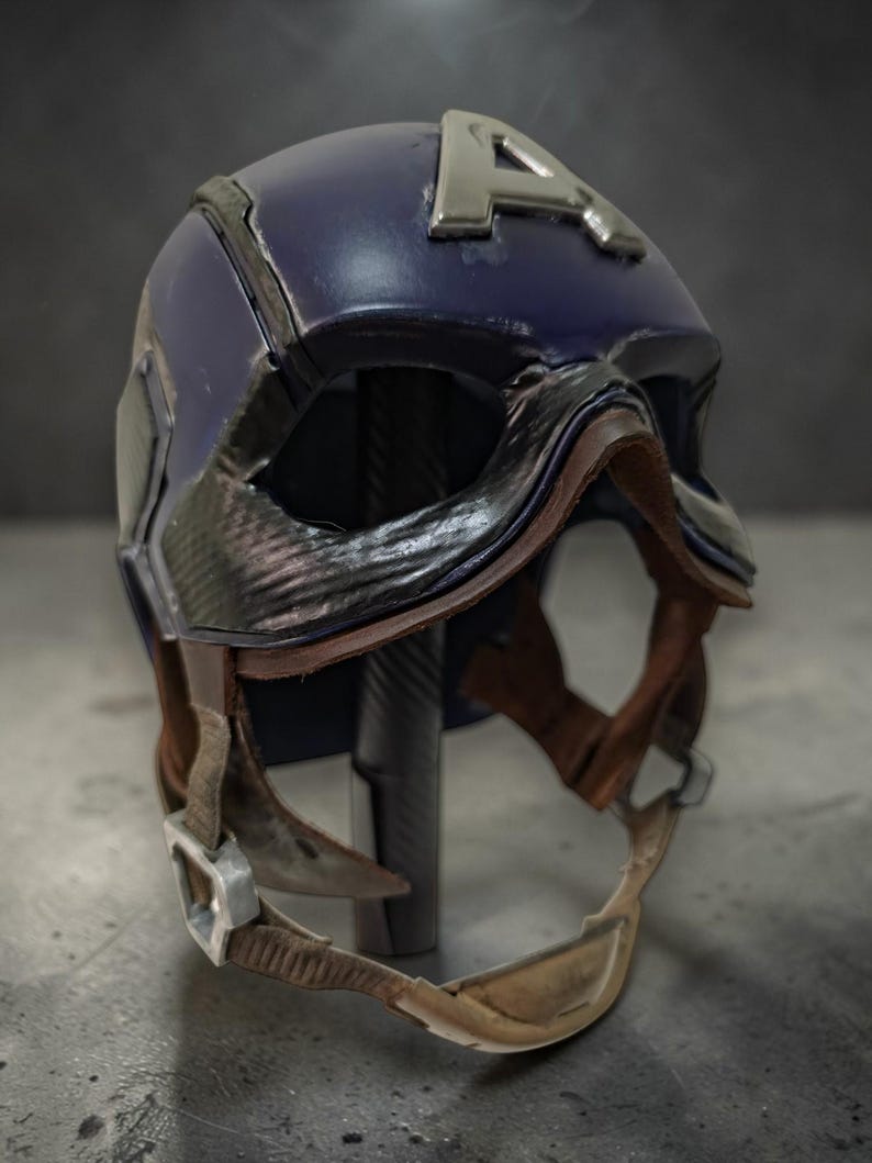 K&ouml;nnte beinhalten: Ein dunkelblauer Helm mit silbernem "A"-Emblem. Der Helm hat ein lederartiges Visier und einen Kinnriemen mit silberner Schnalle. Das Design erinnert an einen Superheldenhelm.