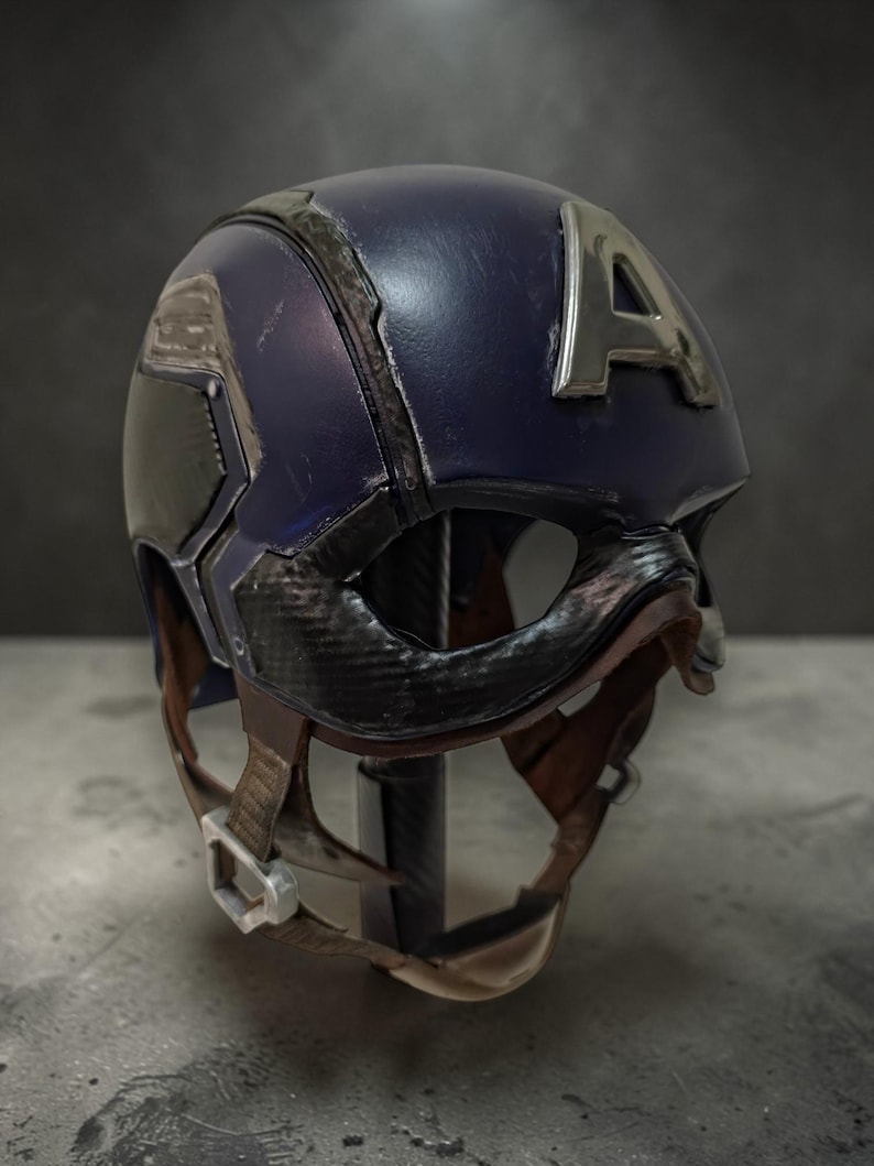 May include: A navy blue Captain America helmet with a silver "A" emblem. The helmet has a black visor and chin strap with a brown leather-like texture. The helmet is displayed on a dark background.