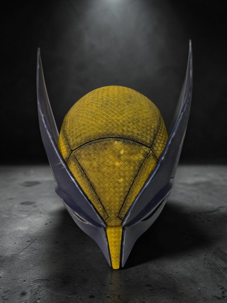 Puede incluir: Una m&aacute;scara inspirada en Wolverine, con una superficie texturizada amarilla y detalles en azul oscuro. La m&aacute;scara tiene orejas puntiagudas y un dise&ntilde;o afilado y angular, sobre un fondo oscuro.