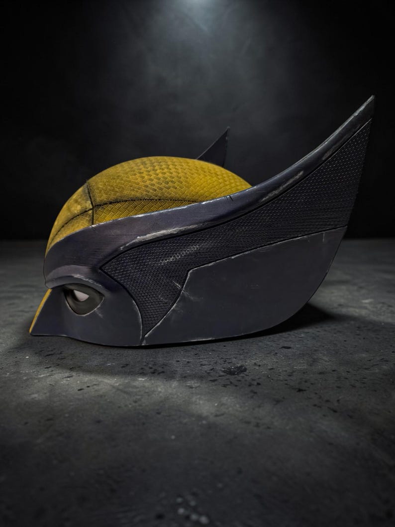 Puede incluir: Primer plano de una m&aacute;scara de Wolverine. La m&aacute;scara presenta una parte superior amarilla texturizada con detalles en negro y orejas puntiagudas. El &aacute;rea de los ojos es negra y la m&aacute;scara est&aacute; sobre un fondo oscuro.