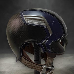 K&ouml;nnte beinhalten: Ein schwarz-blauer Helm mit einer Kohlefaserstruktur auf der linken Seite. Der Helm hat silberne Akzente und einen braunen Kinnriemen mit Metallschnalle. Wahrscheinlich ein Requisit oder Sammlerst&uuml;ck.