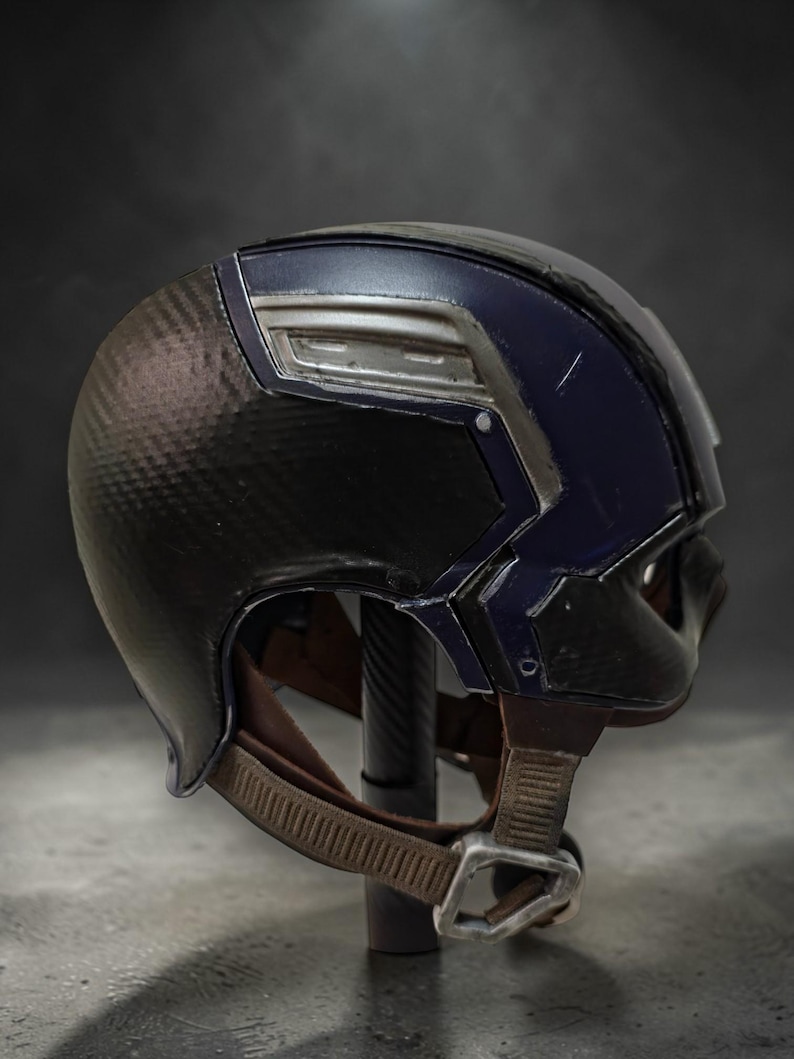 K&ouml;nnte beinhalten: Ein schwarz-blauer Helm mit einer Kohlefaserstruktur auf der linken Seite. Der Helm hat silberne Akzente und einen braunen Kinnriemen mit Metallschnalle. Wahrscheinlich ein Requisit oder Sammlerst&uuml;ck.