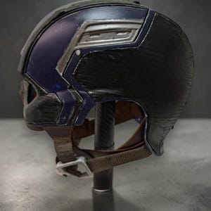 K&ouml;nnte beinhalten: Ein schwarz-blauer Helm mit silbernen Akzenten. Der Helm hat eine strukturierte Oberfl&auml;che und einen braunen Kinnriemen mit silberner Schnalle. Der Helm wird auf einem schwarzen St&auml;nder pr&auml;sentiert.
