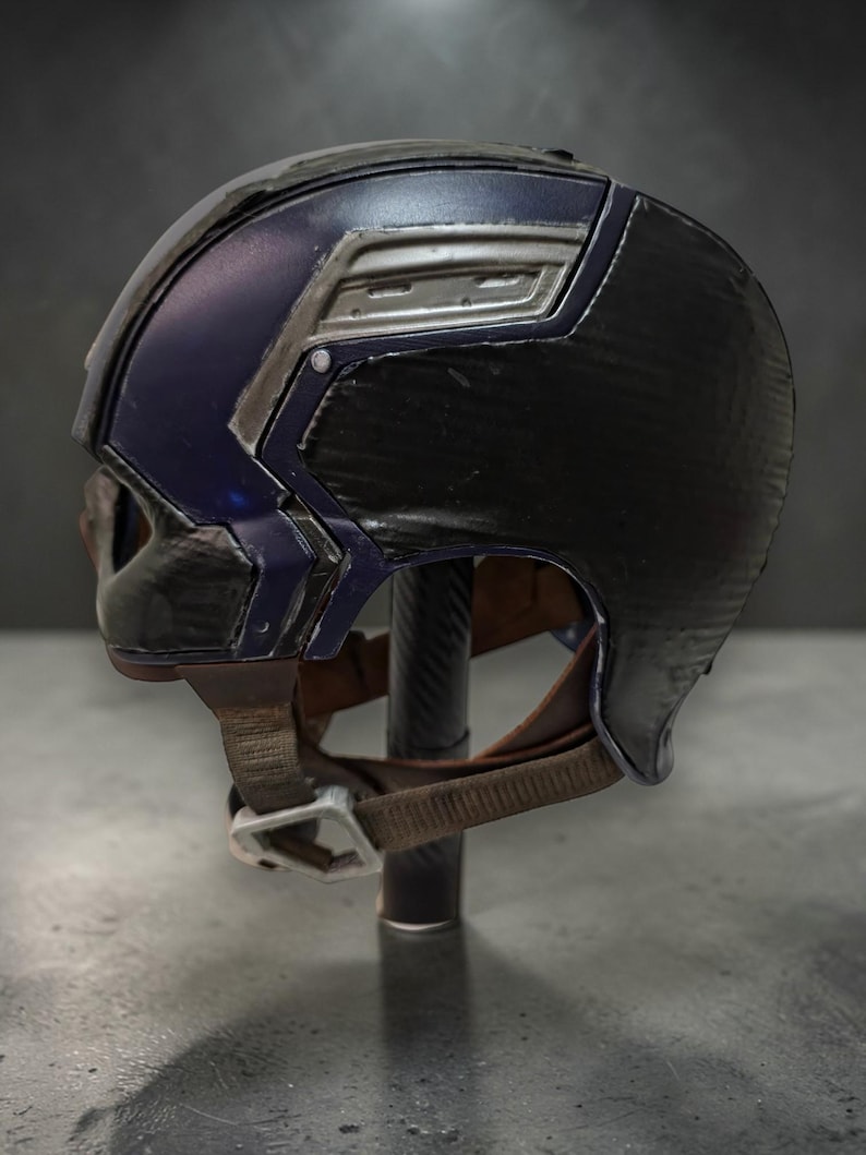 K&ouml;nnte beinhalten: Ein schwarz-blauer Helm mit silbernen Akzenten. Der Helm hat eine strukturierte Oberfl&auml;che und einen braunen Kinnriemen mit silberner Schnalle. Der Helm wird auf einem schwarzen St&auml;nder pr&auml;sentiert.