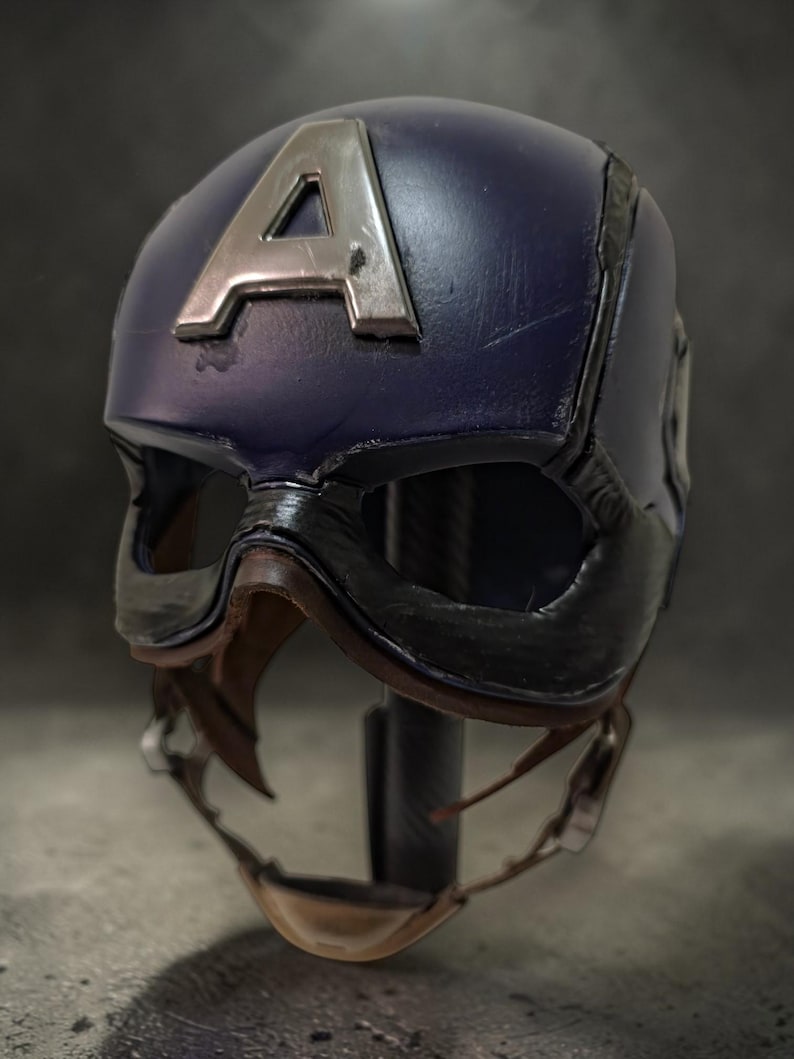 K&ouml;nnte beinhalten: Blauer Captain America Helm mit silbernem "A" auf der Vorderseite. Der Helm hat ein schwarzes Visier und einen Kinnriemen. Eine Nachbildung des Helms der Comicfigur.