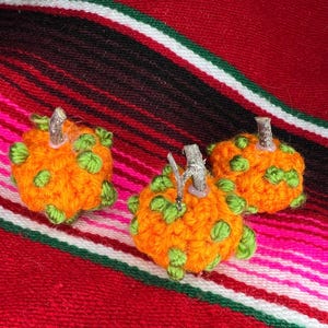 Op de afbeelding: Drie handgemaakte oranje en groene gehaakte pompoenen met kleine houten stelen. De pompoenen zijn gerangschikt op een kleurrijke rood, roze en groen gestreepte textielachtergrond, perfect voor herfstdecoratie.