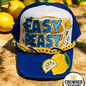 May include: A blue and white trucker hat featuring the text "EASY PEASY" in blue letters over a yellow lemon graphic. The hat includes a yellow braided trim across the front and a yellow lemon slice patch on the blue brim with green leaves.
