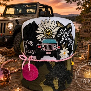 May include: A white and camouflage trucker hat featuring embroidered patches of a daisy, a pink vehicle, and the text "take it easy" and "girl gang". The hat is accented with a pink beaded trim, a pink circular patch, and a small yellow duck charm.