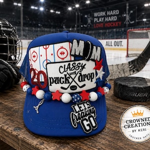 May include: A blue trucker hat featuring a patch with the text 'CLASSY UNTIL PUCK DROP', a 'MOM' patch, a 'LET'S FREAKING GO' patch, and a '20' patch. The hat is decorated with red, white, and blue beads, star-shaped charms, and a hockey stick charm.