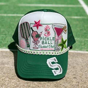 May include: Green and white trucker hat with a curved brim and mesh back. The front panel features embroidered patches of a pickleball paddle, cocktail glasses, and the text "Pickle Ball Cocktail Club". A white and pink rope trim accents the brim, and a large white "S" is on the front.