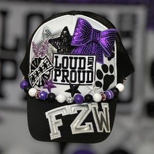 May include: Black and white trucker hat with a white front panel. Embellished with a purple sequin bow, stars, and the text "LOUD AND PROUD". The brim features the letters "FZW" in silver.