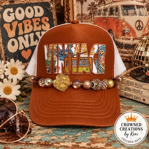 Può includere: Un cappellino trucker color ruggine con retro in rete bianca e pannello frontale con la parola VINTAGE ricamata in un tessuto multicolore a motivi. Un filo decorativo di perline assortite, tra cui un fiore giallo, è posizionato sulla visiera.