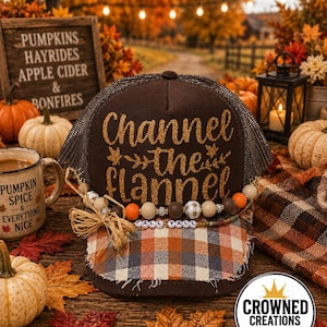 May include: A brown trucker hat with a mesh back and a plaid brim. The front features gold glitter text reading Channel the flannel with leaf accents. A decorative strand of wooden and patterned beads with the text FALL YALL sits above the brim.