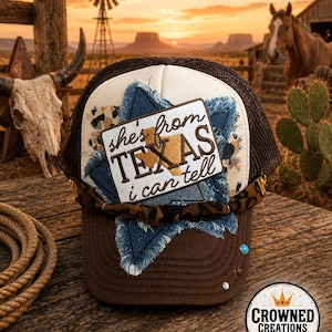 May include: A brown and cream trucker hat featuring a rectangular patch with the text 'she's from TEXAS i can tell' and a gold-colored Texas state outline. The hat includes a frayed blue denim star, a leopard print band, and small studs.