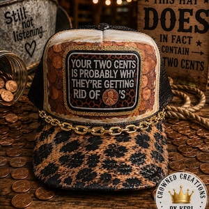 May include: A trucker hat featuring a leopard print brim and a front panel decorated with a pattern of copper pennies. A central rectangular patch displays the text: YOUR TWO CENTS IS PROBABLY WHY THEY'RE GETTING RID OF 'S, with a real penny attached.