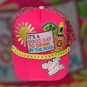 Könnte beinhalten: Leuchtend pinkfarbene Trucker-Cap mit Mesh-Rückseite und gebogenem Schirm. Die Cap hat aufgestickte Aufnäher von Sonne, Limette, Tequila-Flasche und Text mit der Aufschrift "It's a good day to drink by the pool" und "Girls just wanna have fun."