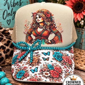 May include: A beige trucker hat featuring a graphic illustration of a woman with long wavy hair and a floral outfit. The brim is decorated with a floral pattern in shades of red and teal, accented by a teal beaded string tied into a bow.