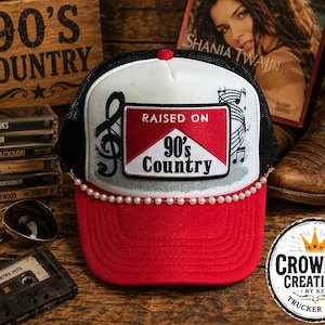 Può includere: Un cappellino trucker rosso e bianco con retro in rete nera e visiera rossa. Una toppa rettangolare sul davanti mostra la scritta Raised on 90's Country con una nota musicale. Una fila di piccole perle bianche decora la base del pannello frontale.