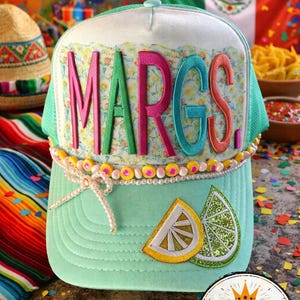 May include: A mint green trucker hat with the word "MARGS" in colorful embroidered letters. The hat features a floral pattern, lemon slice patches, and a beaded accent. The hat is decorated with a Cinco de Mayo theme.