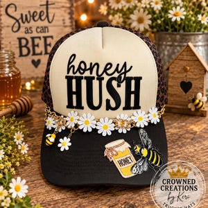 May include: A black and cream trucker hat featuring the text honey HUSH in black lettering. The hat is decorated with a gold chain, white daisy appliques, a small bee charm, and a patch depicting a honey jar with a bee on the black brim.