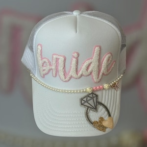 May include: White trucker hat with "bride" in pink script. The hat features a pearl and floral embellishment, and a ring charm with a diamond and gold heart. The mesh back is white.