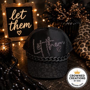 May include: A black trucker hat featuring a mesh back and a brim with a subtle leopard print pattern. The front panel displays the text "let them" in silver rhinestones, accented by a thick black chain detail across the base of the crown.
