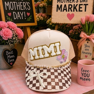 May include: A beige and brown trucker hat with the name "MIMI" in gold letters and a pink flower. The hat has a checkered pattern on the brim and a pearl and gold chain. A pink mug with "love YOU mom" is also visible.