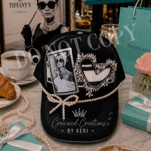 May include: A black baseball cap featuring a rectangular black and white photograph of a person, a heart-shaped rhinestone embellishment, a pearl-beaded bow, and the white printed text Crowned Creations BY KERI with a crown icon above it.