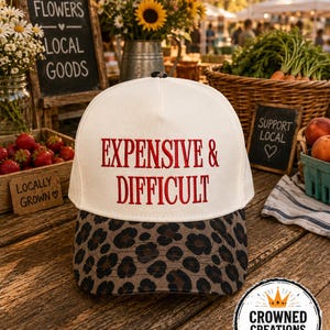 Könnte beinhalten: Eine weiße Trucker-Cap mit dem in Rot gestickten Text EXPENSIVE & DIFFICULT auf der Vorderseite. Die Kappe hat einen gebogenen Schirm mit einem braun-schwarzen Leopardenmuster. Das Design umfasst einen Knopf oben in der Mitte der Krone.
