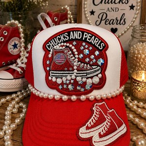 May include: A red and white trucker hat featuring a central patch with the text "CHUCKS AND PEARLS" and an embroidered red sneaker with an American flag detail. The hat is accented with a string of white pearls and a secondary patch of two red sneakers.